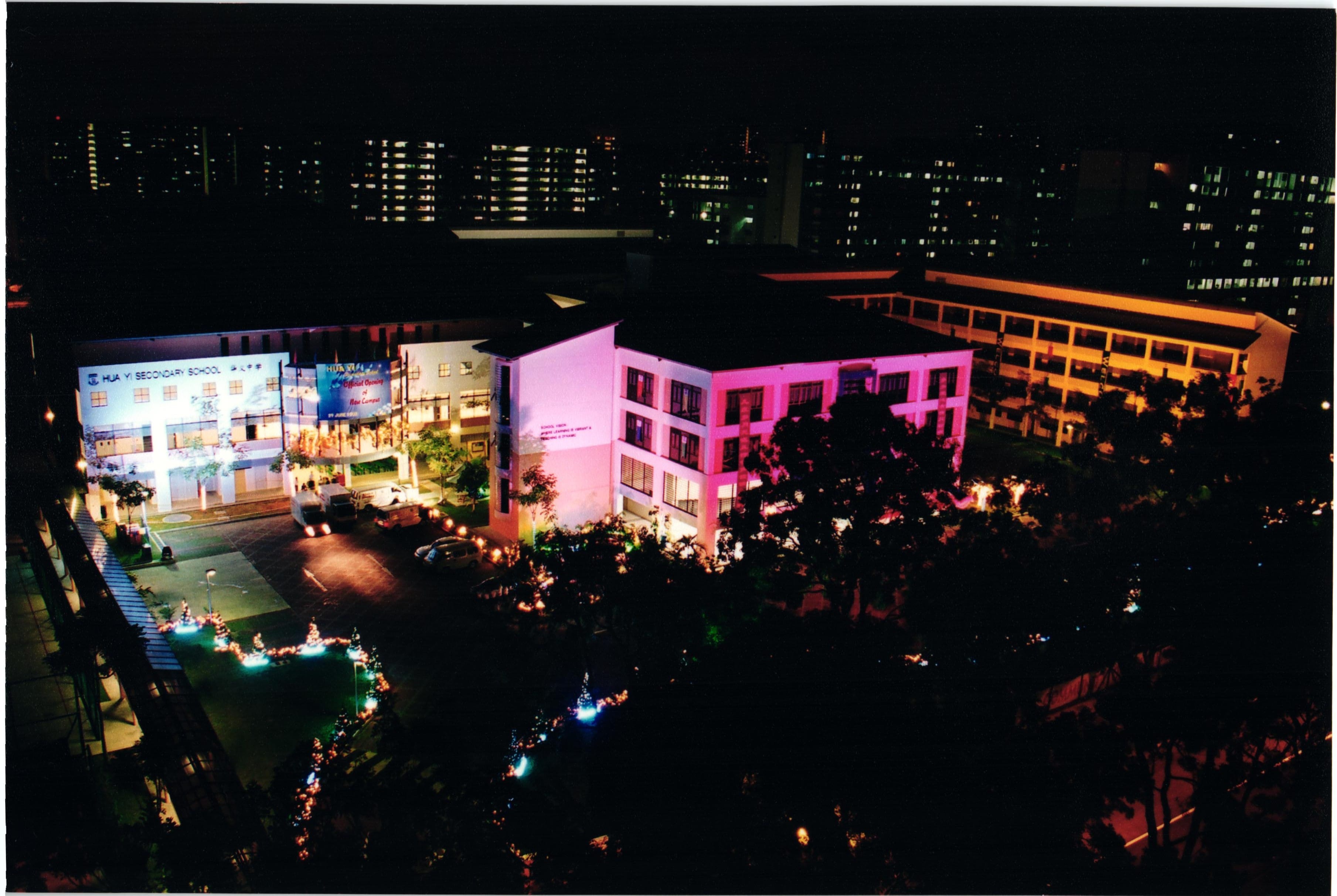 Aerial night view of Hua Yi Secondary School campus lit with pink and blue lights, with city buildings in background.